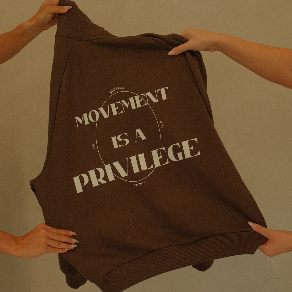 Brown hoodie with 'Movement is a Privilege' text held by hands against a beige background