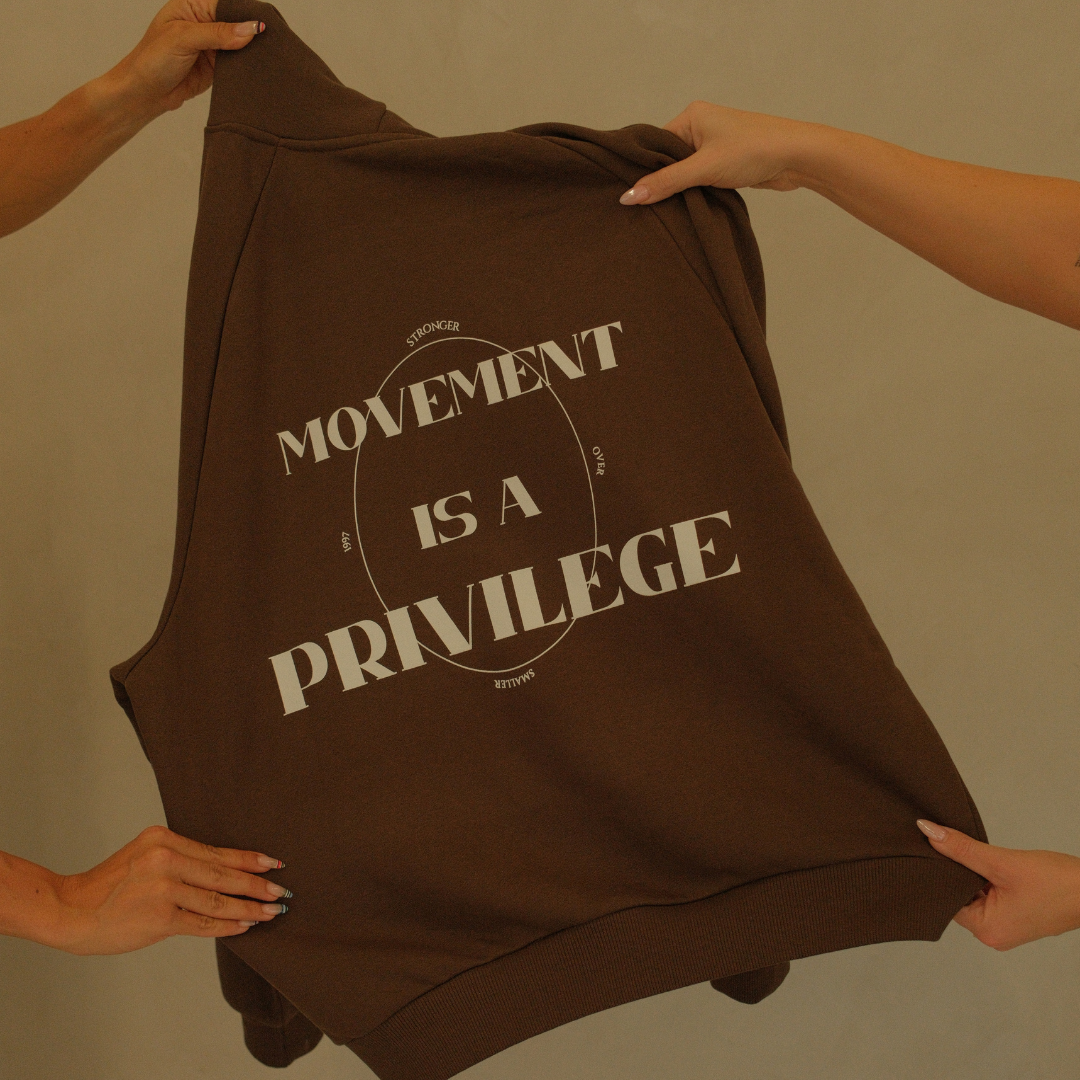 Brown hoodie with 'Movement is a Privilege' text held by hands against a beige background